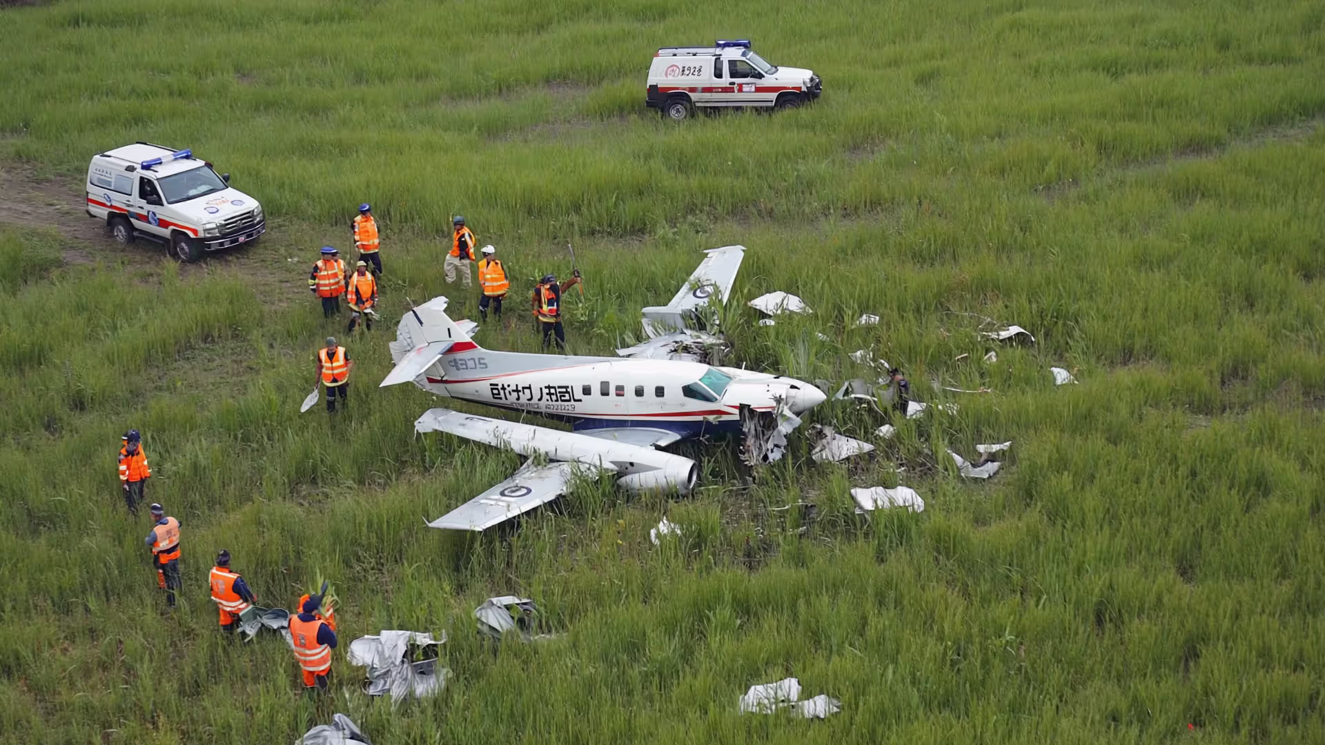 Aerial view of a small plane crash site in a green field with emergency responders and rescue vehicles